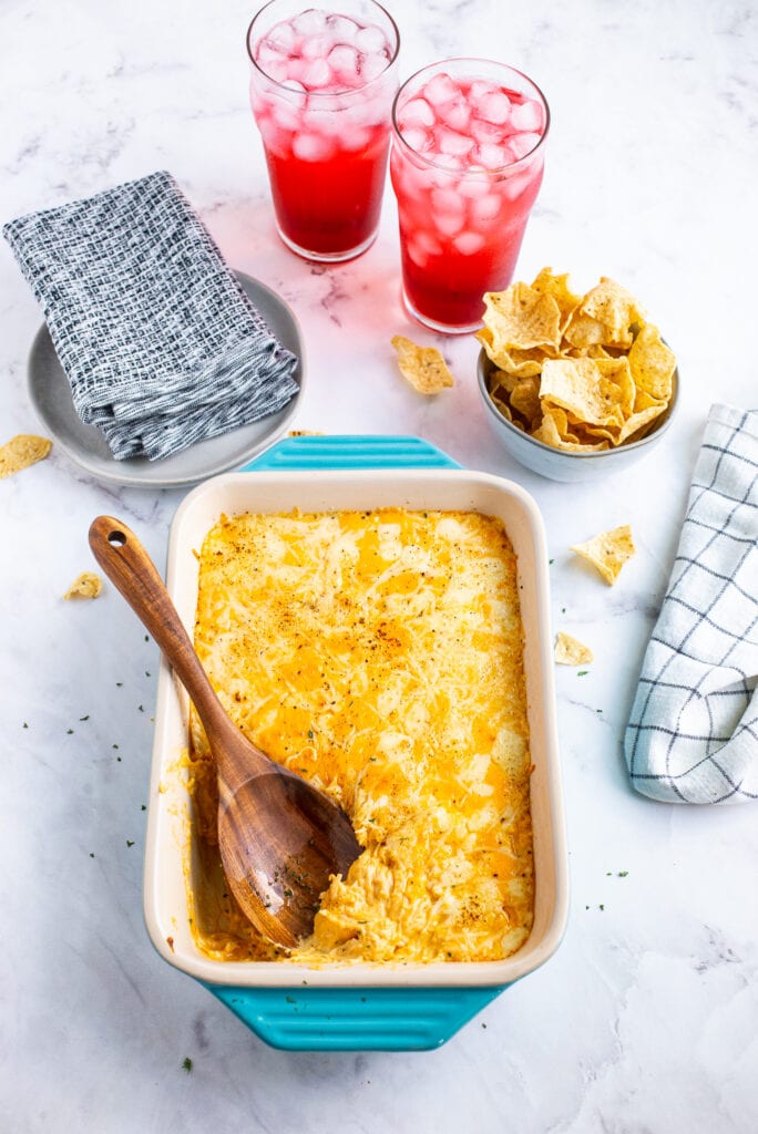 Casserole dish of Buffalo Chicken dip with a wooden spoon, drinks, chips, and plates and napkins.