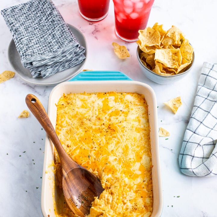 Buffalo Chicken Dip in a blue baking dish with a wooden spoon surrounded by a bowl of chips and drinks