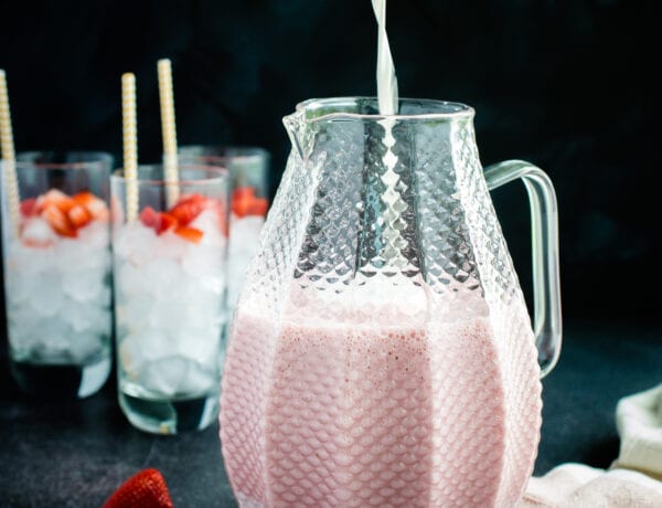Milk being poured into a pitcher with a blended strawberry mitxture. Glasses of ice with strawberries are located in the background.